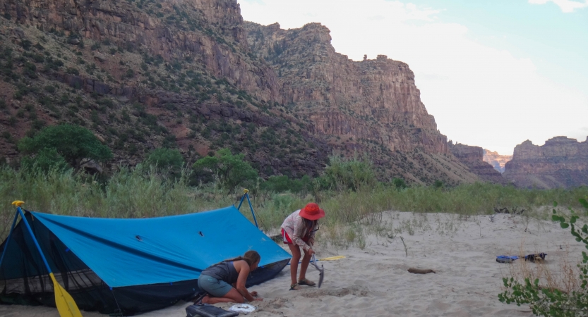 Two people set up a tarp shelter in the sand. In the background, tall canyon walls reach toward a blue sky.